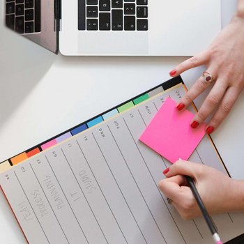 Ein Notizbuch und ein pinker Zettel liegen neben einem Laptop. Eine Person schreibt etwas auf den Zettel.